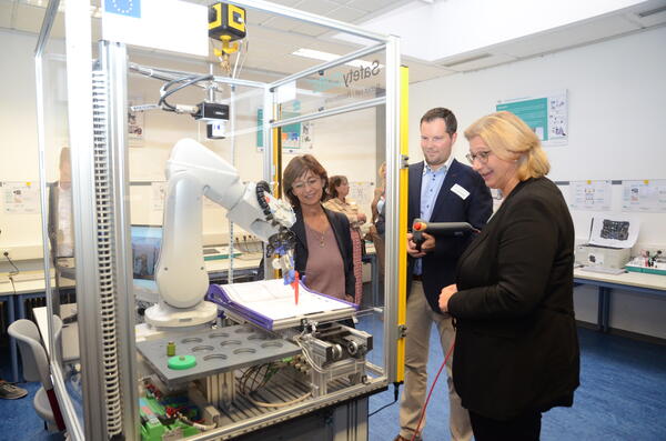 Lehrer Alexander Bohlen zeigt Ministerpräsidentin Anke Rehlinger und Landrätin Daniela Schlegel-Friedrich, wie die Auszubildenden im neuen Kompetenzzentrum die Bedienung eines Sechs-Achs-Roboters erlernen.